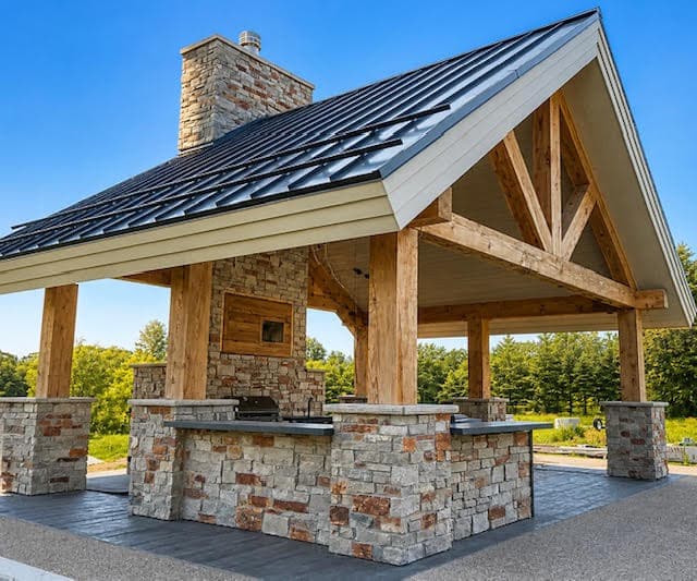 Timber frame outdoor pavilion with a stone fireplace, kitchen, and dark metal gabled roof.